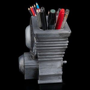 May include: A gray, metallic-finished pencil holder shaped like a mechanical engine block with cooling fins. The container is filled with various colored pencils, black pens, a red pen, and a clear-barreled pen, all standing upright in the central cavity.