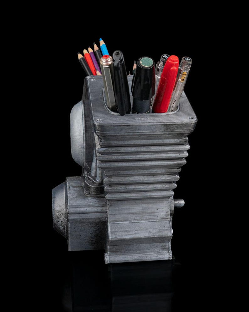 May include: A gray, metallic-finished pencil holder shaped like a mechanical engine block with cooling fins. The container is filled with various colored pencils, black pens, a red pen, and a clear-barreled pen, all standing upright in the central cavity.