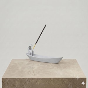May include: A matte white ceramic incense holder shaped like a small boat with a seated figure wearing a wide-brimmed hat. The figure holds a thin, black incense stick with a yellow base, which extends upward from the center of the boat.