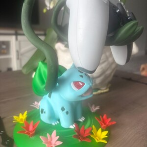 Op de afbeelding: Een lichtblauw Bulbasaur-figuur met een groene bol op de rug, die dient als standaard voor een wit-zwarte videogamecontroller. Het figuur staat op een ronde groene basis versierd met kleine rode, gele en roze bloemaccenten.