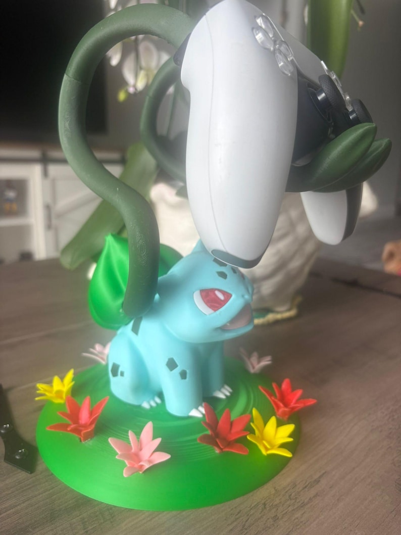 Op de afbeelding: Een lichtblauw Bulbasaur-figuur met een groene bol op de rug, die dient als standaard voor een wit-zwarte videogamecontroller. Het figuur staat op een ronde groene basis versierd met kleine rode, gele en roze bloemaccenten.