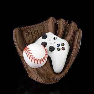 Puede incluir: Un guante de b&eacute;isbol de cuero marr&oacute;n que sostiene una pelota de b&eacute;isbol blanca con costuras rojas y un mando de videojuego inal&aacute;mbrico blanco con joysticks negros, una cruceta negra y cuatro botones de acci&oacute;n de colores A, B, X e Y.