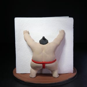 May include: A napkin holder featuring a beige sumo wrestler figure with a black topknot and a red mawashi belt. The figure is positioned on a round brown base, with arms raised to hold a stack of square white paper napkins in place.