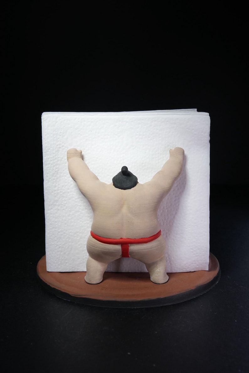 May include: A napkin holder featuring a beige sumo wrestler figure with a black topknot and a red mawashi belt. The figure is positioned on a round brown base, with arms raised to hold a stack of square white paper napkins in place.