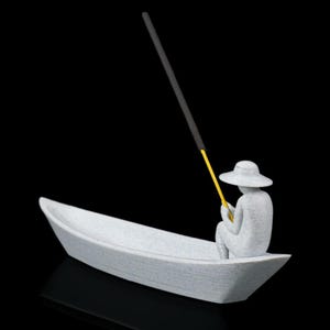 May include: A light gray textured figurine of a person wearing a wide-brimmed hat, seated inside a small boat. The figure holds a thin yellow rod that extends upward, supporting a long, dark gray incense stick angled toward the sky.
