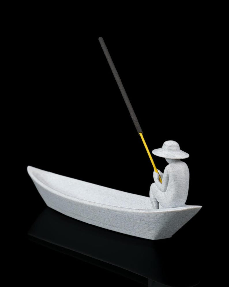 May include: A light gray textured figurine of a person wearing a wide-brimmed hat, seated inside a small boat. The figure holds a thin yellow rod that extends upward, supporting a long, dark gray incense stick angled toward the sky.