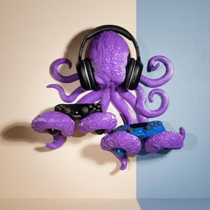 Könnte beinhalten: Eine lila Wandskulptur in Form eines Oktopus mit texturierten Tentakeln, die dazu dienen, Gegenstände zu halten. Der Oktopus trägt einen schwarzen Over-Ear-Kopfhörer auf dem Kopf und hält zwei Gamecontroller, einen schwarzen und einen blauen.