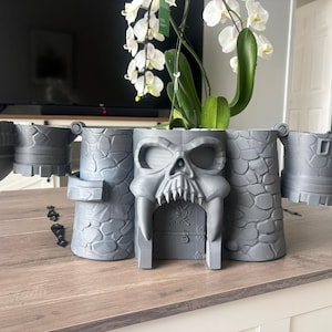 Op de afbeelding: Een grijze 3D-geprinte plantenbak in kasteelthema met een opvallend schedelontwerp bij de centrale ingang. De structuur bevat muren met een steenachtige textuur, zijtorens en scharnierende delen die naar buiten uitsteken voor een brede uitstraling.