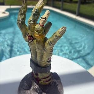 May include: A sculpted figurine of a decaying, yellowish-green zombie hand emerging from a dark, textured base. The hand features visible stitches, a gold-colored ring on the ring finger, and a dark grey cuff around the wrist, with red accents.
