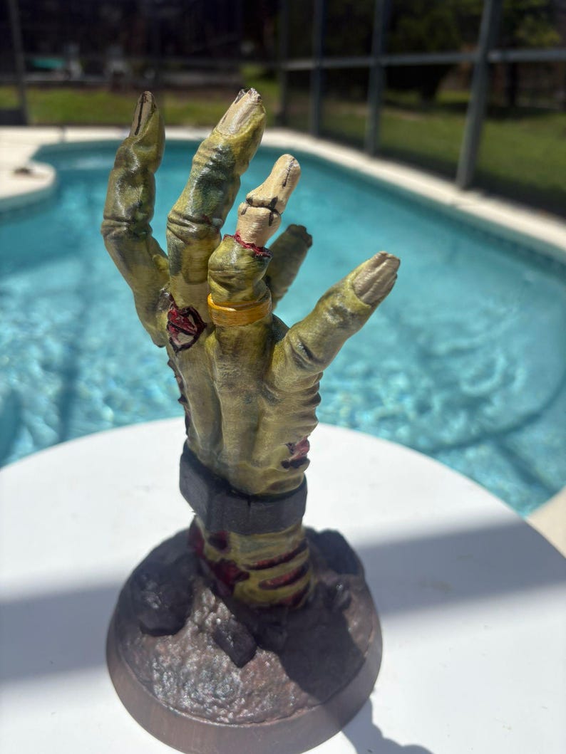 May include: A sculpted figurine of a decaying, yellowish-green zombie hand emerging from a dark, textured base. The hand features visible stitches, a gold-colored ring on the ring finger, and a dark grey cuff around the wrist, with red accents.