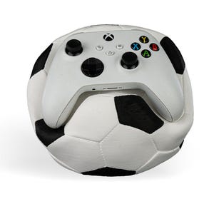 Op de afbeelding: Een witte draadloze videogamecontroller met zwarte joysticks en veelkleurige actieknoppen die op een zwart-witte voetbal rust. De controller is voorzien van het Xbox-logo in het midden bovenaan en standaard directionele invoeropties.