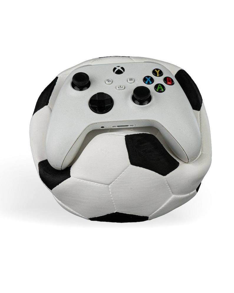 Op de afbeelding: Een witte draadloze videogamecontroller met zwarte joysticks en veelkleurige actieknoppen die op een zwart-witte voetbal rust. De controller is voorzien van het Xbox-logo in het midden bovenaan en standaard directionele invoeropties.