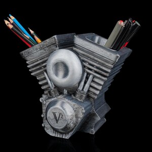 May include: A metallic gray V-twin engine shaped desk organizer featuring two cylindrical compartments for holding pens and pencils. The front displays a circular V2 emblem with a weathered finish, and the structure includes detailed cooling fins.