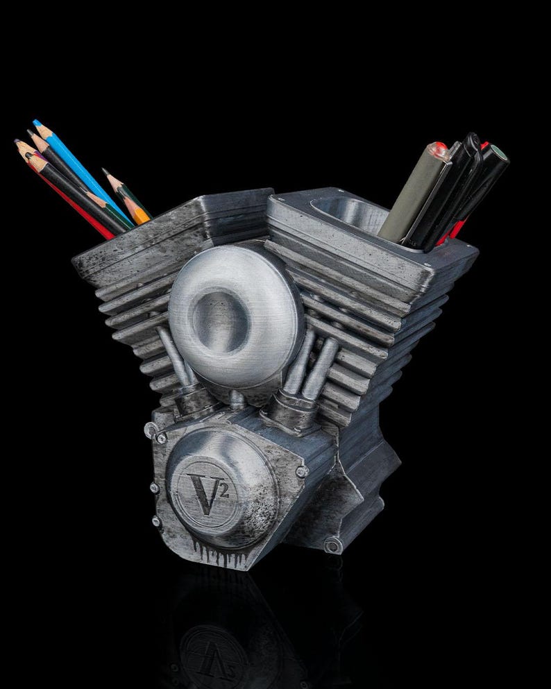 May include: A metallic gray V-twin engine shaped desk organizer featuring two cylindrical compartments for holding pens and pencils. The front displays a circular V2 emblem with a weathered finish, and the structure includes detailed cooling fins.
