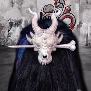 May include: A hair accessory featuring a pale, textured dragon skull with two prominent curved horns and smaller side spikes. A long, bone-shaped pin extends horizontally through the skull, securing it against a mass of dark, straight hair.