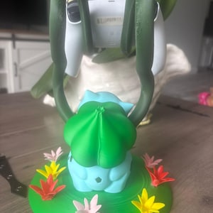 Op de afbeelding: Een lichtblauwe en groene controllerstandaard in de vorm van een wezen met twee lange, donkergroene, rankachtige armen die omhoog reiken om een witte controller vast te houden. De basis is een rond groen platform versierd met kleine, kleurrijke bloemen.