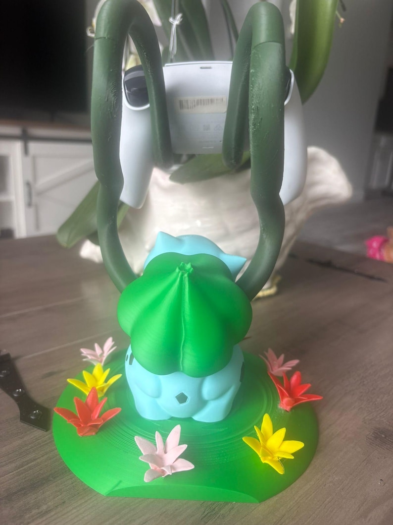 Op de afbeelding: Een lichtblauwe en groene controllerstandaard in de vorm van een wezen met twee lange, donkergroene, rankachtige armen die omhoog reiken om een witte controller vast te houden. De basis is een rond groen platform versierd met kleine, kleurrijke bloemen.