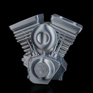 May include: A silver-colored V-twin engine model featuring prominent cooling fins on the cylinder heads and a circular side cover embossed with the text V2. The metallic structure displays visible horizontal layer lines from the manufacturing process.