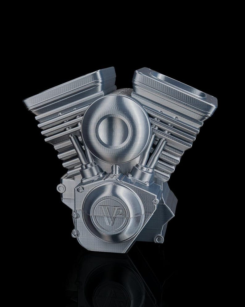 May include: A silver-colored V-twin engine model featuring prominent cooling fins on the cylinder heads and a circular side cover embossed with the text V2. The metallic structure displays visible horizontal layer lines from the manufacturing process.
