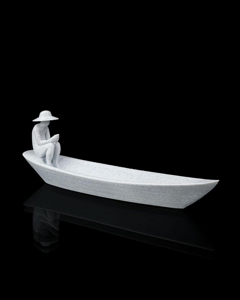 May include: A white, textured sculpture of a figure wearing a wide-brimmed hat, sitting inside a long, narrow boat. The figure is positioned at one end of the vessel, with knees bent and hands held together, appearing to look down into the boat.