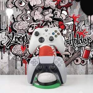 May include: A white Xbox controller rests on top of a red and white sneaker-shaped stand, which holds a white and black PlayStation controller in its front section. The stand is mounted on a circular green base, designed for gaming controller storage.