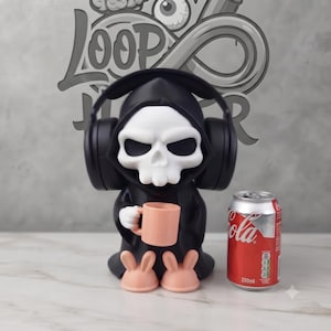 May include: A black and white skull figure wearing black over-ear headphones, holding a light pink mug, and wearing light pink bunny slippers. A 330ml Coca-Cola can is placed next to the figure for scale, showing the relative size of the object.