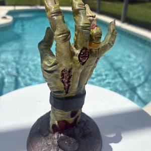 May include: A sculpted hand figurine featuring a weathered, olive-green skin texture with visible red lacerations and exposed tissue. The hand wears a dark gray wristband and a gold-colored ring on the ring finger, mounted on a circular base.