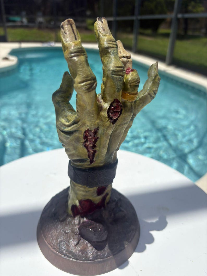 May include: A sculpted hand figurine featuring a weathered, olive-green skin texture with visible red lacerations and exposed tissue. The hand wears a dark gray wristband and a gold-colored ring on the ring finger, mounted on a circular base.