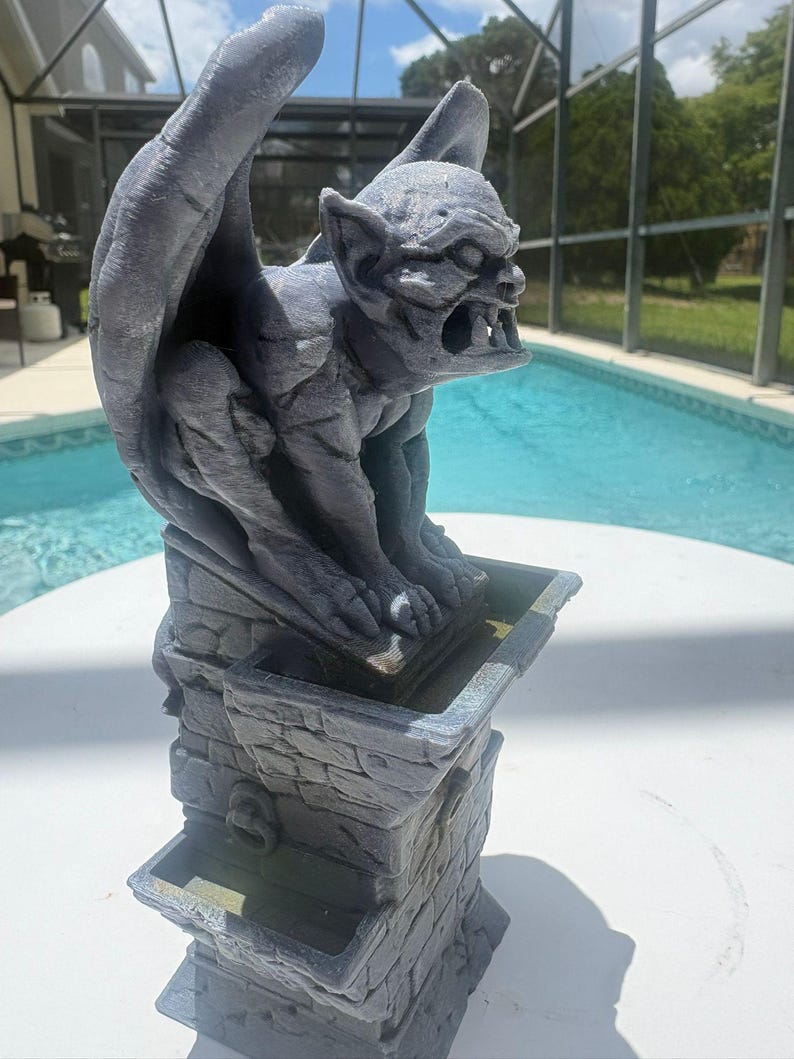 Puede incluir: Una estatua de g&aacute;rgola gris posada sobre un pedestal con textura de piedra. La g&aacute;rgola presenta alas grandes, orejas puntiagudas y una boca abierta con dientes visibles. El pedestal tiene una cuenca rectangular en la parte superior.