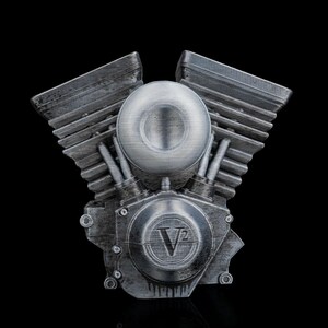 May include: A silver-toned V-twin engine model featuring a weathered metallic finish with prominent cooling fins on the cylinder heads. The central circular housing displays the embossed text V2, and the overall design replicates the mechanical structure of a motorcycle engine with a textured, industrial appearance.