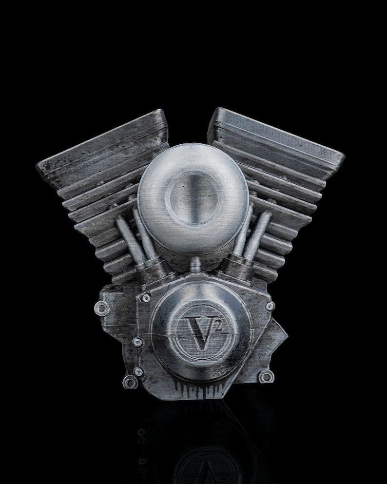 May include: A silver-toned V-twin engine model featuring a weathered metallic finish with prominent cooling fins on the cylinder heads. The central circular housing displays the embossed text V2, and the overall design replicates the mechanical structure of a motorcycle engine with a textured, industrial appearance.