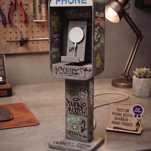 Peut inclure: Un support de chargeur en forme de cabine téléphonique grise, orné de graffitis et d'autocollants. Le mot PHONE est inscrit en bleu en haut. Un socle de charge blanc circulaire est fixé au centre. Une pancarte indique CUSTOM PHONE BOOTH CHARGER - HAND-TAGGED.