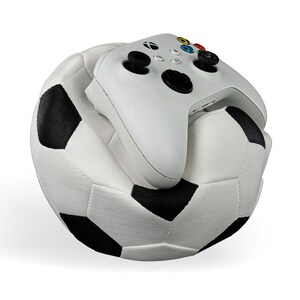 Op de afbeelding: Een witte videogamecontroller met zwarte joysticks en gekleurde knoppen die op een pluche voetbal ligt met een klassiek wit en zwart vijfhoekig patroon. De controller is voorzien van het Xbox-logo en standaard directionele invoer.
