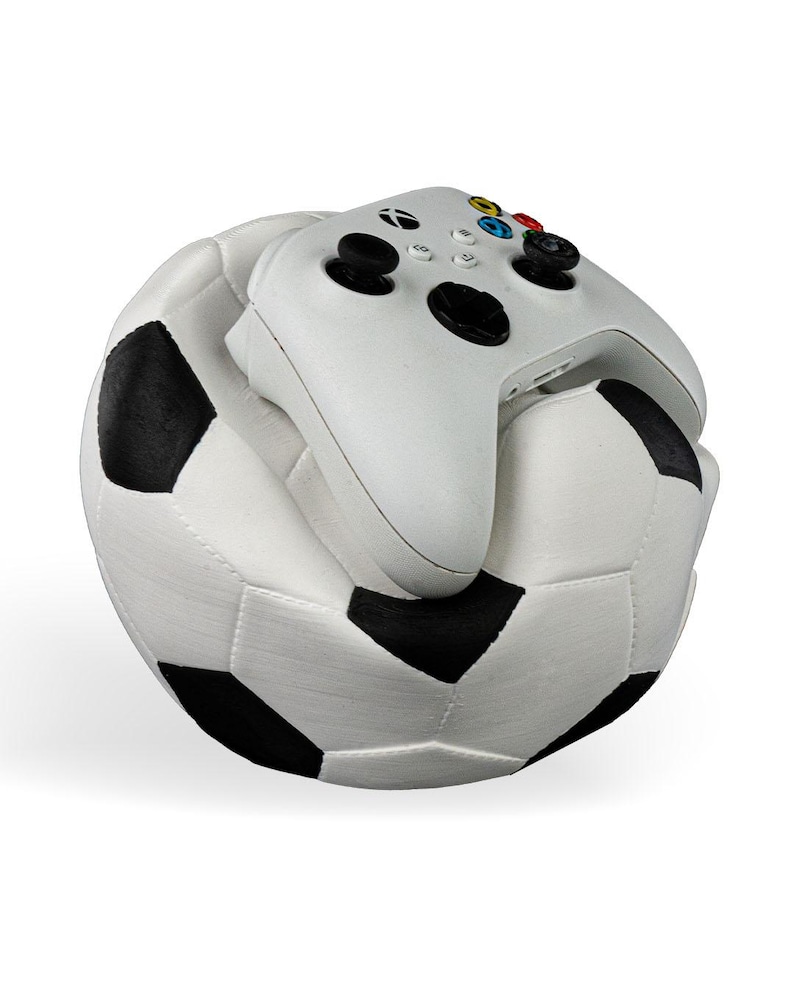 Op de afbeelding: Een witte videogamecontroller met zwarte joysticks en gekleurde knoppen die op een pluche voetbal ligt met een klassiek wit en zwart vijfhoekig patroon. De controller is voorzien van het Xbox-logo en standaard directionele invoer.