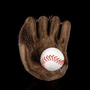 Puede incluir: Un guante de b&eacute;isbol marr&oacute;n con superficie texturizada que sostiene una pelota de b&eacute;isbol blanca con costuras rojas prominentes. El guante tiene compartimentos para los dedos y un dise&ntilde;o de red, sobre un fondo negro con un sutil reflejo.