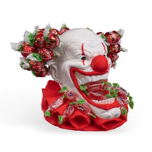 May include: A white clown head sculpture with a red nose and red mouth, featuring a red ruffled collar. The clown is adorned with red-wrapped sweets attached to the hair area and spilling from the open mouth, with the text Showpop on the wrappers.