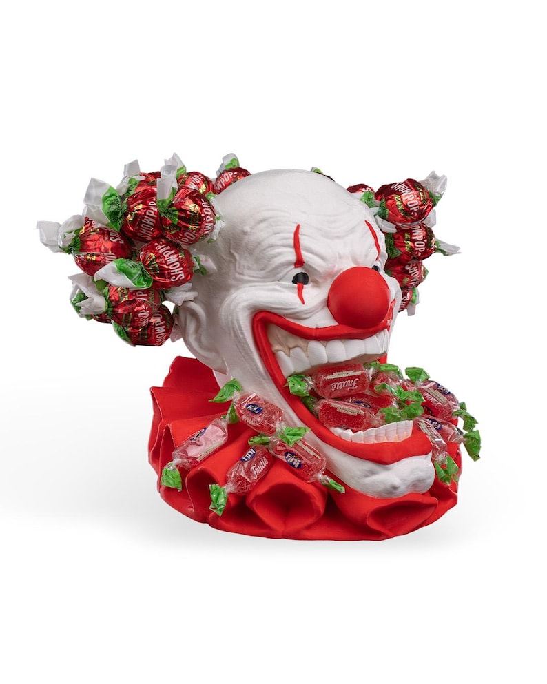 May include: A white clown head sculpture with a red nose and red mouth, featuring a red ruffled collar. The clown is adorned with red-wrapped sweets attached to the hair area and spilling from the open mouth, with the text Showpop on the wrappers.