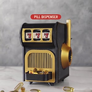 May include: A black and gold Pill-O-Matic pill dispenser shaped like a slot machine. The device features three windows displaying the number 7 in red, a vertical lever on the side, and a small tray at the bottom containing several yellow capsules.