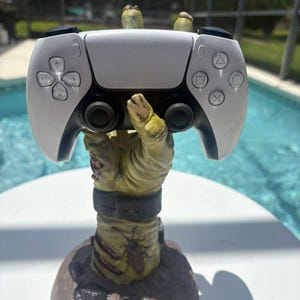 May include: A white and black video game controller resting on a sculpted, yellowish-green zombie hand stand. The hand features a dark wristband, a small insect graphic, and textured skin details, rising from a circular base with a rocky appearance.