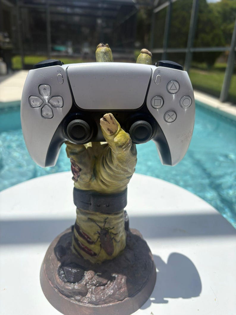 May include: A white and black video game controller resting on a sculpted, yellowish-green zombie hand stand. The hand features a dark wristband, a small insect graphic, and textured skin details, rising from a circular base with a rocky appearance.
