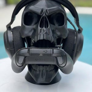 May include: A matte black 3D printed skull headphone stand featuring a realistic anatomical design. The skull is wearing a pair of black over-ear gaming headphones and is holding a black wireless gaming controller within its open jaw.