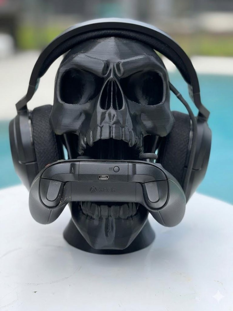 May include: A matte black 3D printed skull headphone stand featuring a realistic anatomical design. The skull is wearing a pair of black over-ear gaming headphones and is holding a black wireless gaming controller within its open jaw.
