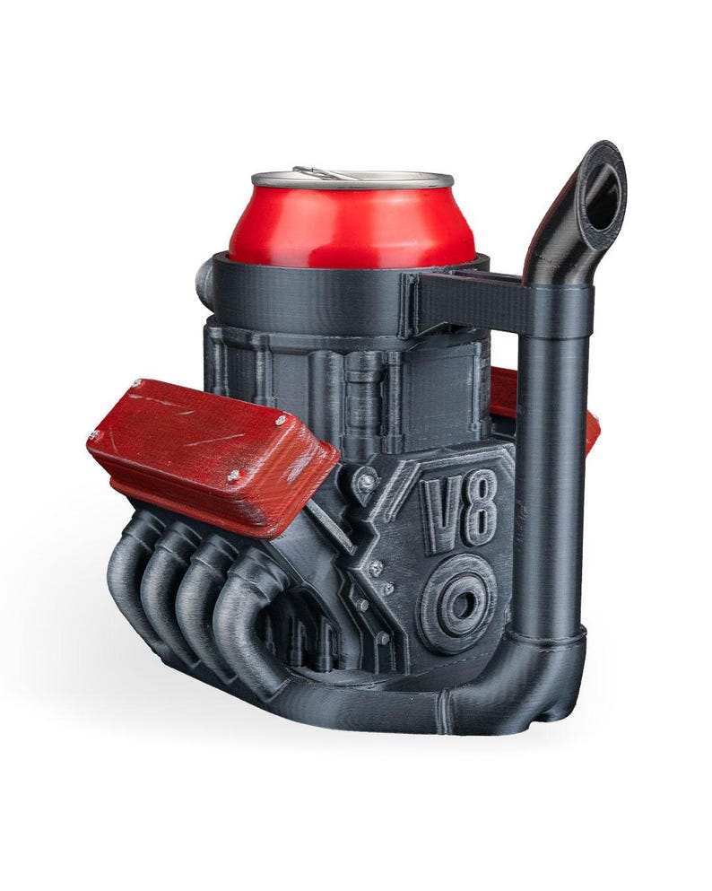 May include: A black and red V8 engine-shaped beverage can holder featuring a prominent exhaust pipe handle. The structure is molded with detailed engine components, including red valve covers and the text V8 embossed on the side, designed to securely hold a standard red aluminum drink can.