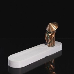May include: A bronze-toned figurine of a person wearing a conical hat and flowing robes, positioned on one end of an elongated, white, oval-shaped tray with a recessed center section. The object is set against a solid black background.