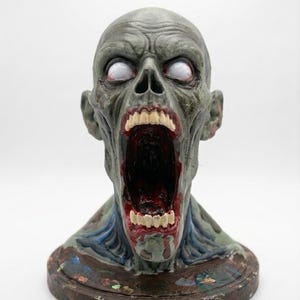 May include: A sculpted bust of a humanoid creature with grey skin, a wide-open mouth revealing dark red interior details, and white, pupil-less eyes. The figure features prominent facial muscles, visible teeth, and sits on a circular base.