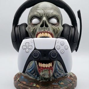 May include: A gray zombie head bust with white eyes and an open mouth holding a white and black video game controller. The zombie head is wearing black over-ear gaming headphones with a microphone boom arm attached to the side of the headset.