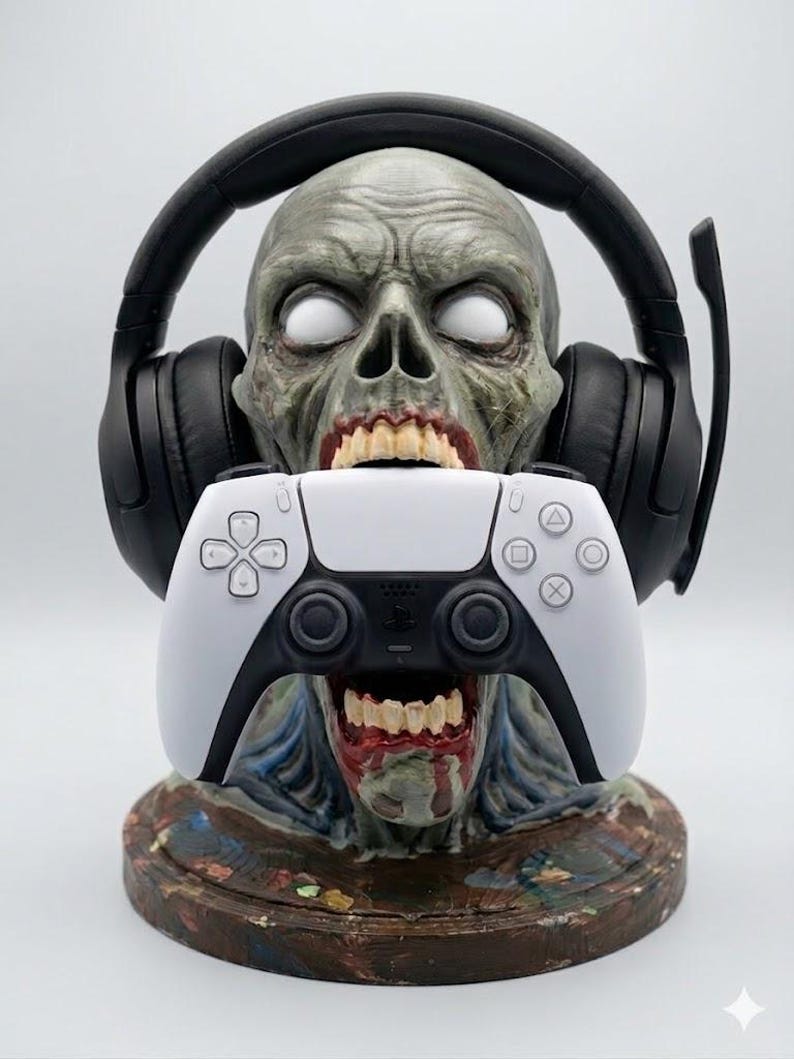 May include: A gray zombie head bust with white eyes and an open mouth holding a white and black video game controller. The zombie head is wearing black over-ear gaming headphones with a microphone boom arm attached to the side of the headset.