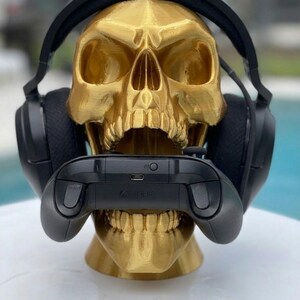 May include: A metallic gold-colored human skull sculpture serving as a stand, featuring black over-ear headphones resting on the cranium and a black wireless gaming controller held horizontally within the open jaw of the skull structure.