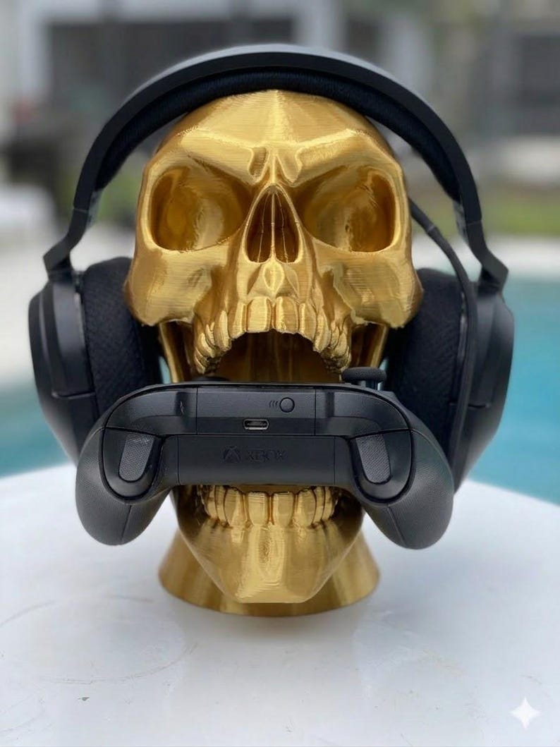 May include: A metallic gold-colored human skull sculpture serving as a stand, featuring black over-ear headphones resting on the cranium and a black wireless gaming controller held horizontally within the open jaw of the skull structure.