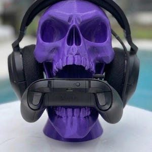 May include: A purple 3D printed skull display stand holding a pair of black over-ear gaming headphones on its cranium and a black wireless gaming controller positioned horizontally within its open jaw, showcasing a matte finish and textured surface.
